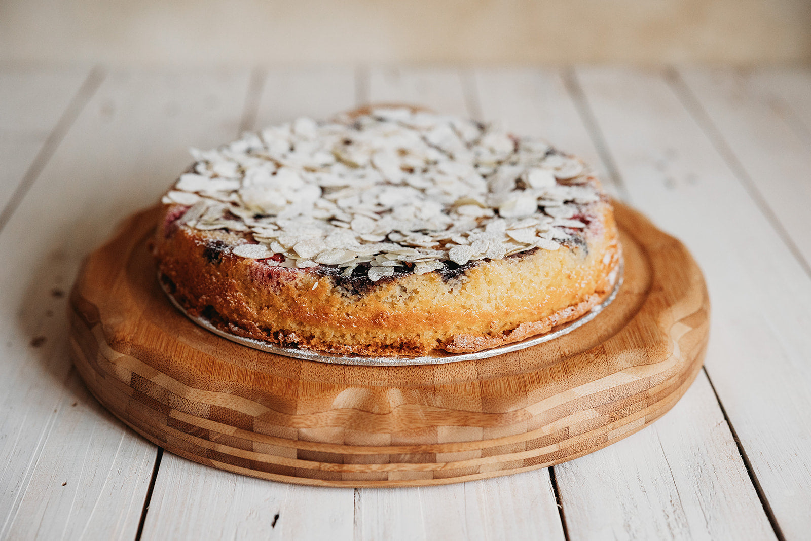 A round almond cake with cherry and berry filling, topped with chopped almonds, served on a wooden board with fresh berries on the side. Handmade in Cambridge by Biscotti di Debora.