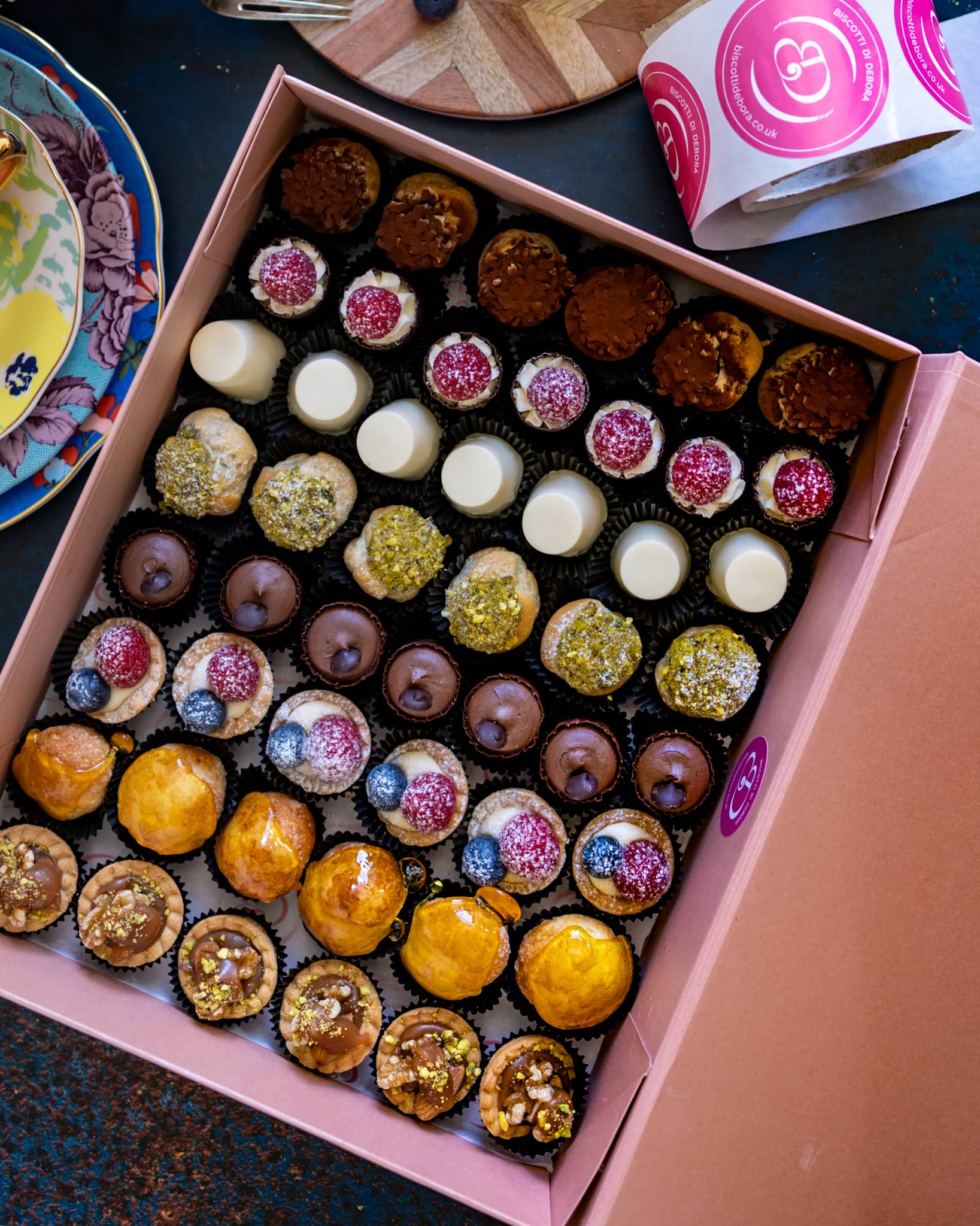 An assortment of petite desserts in a box, featuring an assortment of mini desserts, like pannacottas, choux buns filled with crème patisserie, mini meringues sandwiched with cream, shortcrust tartlets with strawberries. Handmade in Cambridge by Biscotti di Debora.