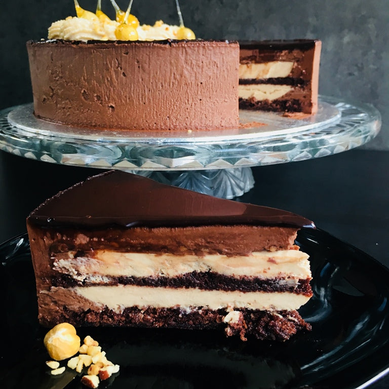 A slice of Torta Setteveli chocolate cake with hazelnut mousse and chocolate sponge layers on a plate, with the whole cake visible in the background. Made by Italian bakery Biscotti di Debora of Cambridge.