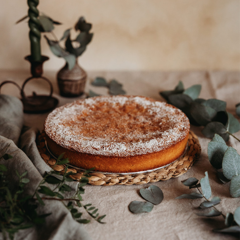 Round coconut and almond cake with powdered sugar on a woven tray, surrounded by greenery