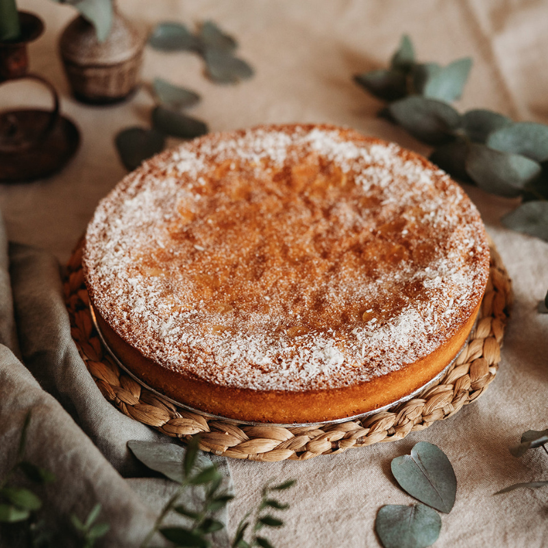 Round cake with powdered sugar on a woven plate, surrounded by green leaves on a neutral background