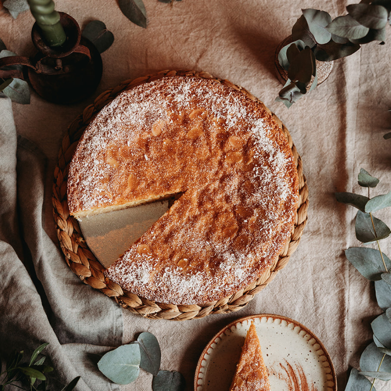 Round cake with a slice removed, dusted with powdered sugar, on a woven tray with a beige cloth background.