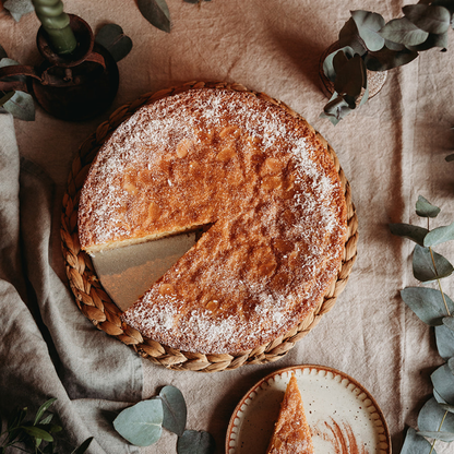 Round cake with a slice removed, dusted with powdered sugar, on a woven tray with a beige cloth background.