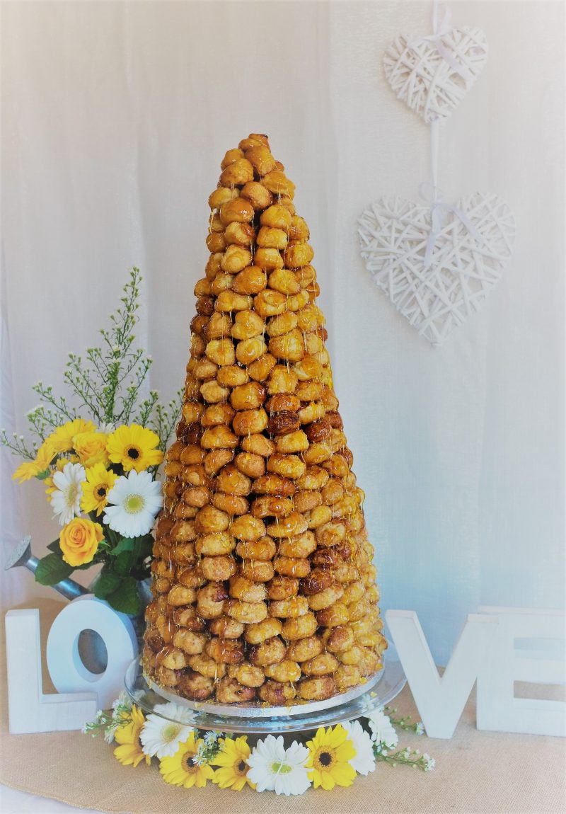 A tall, tiered croquembouche made of profiteroles, filled with cream, and coated in caramel, standing on a plate with decorative flowers and a white heart-shaped sign in the background.