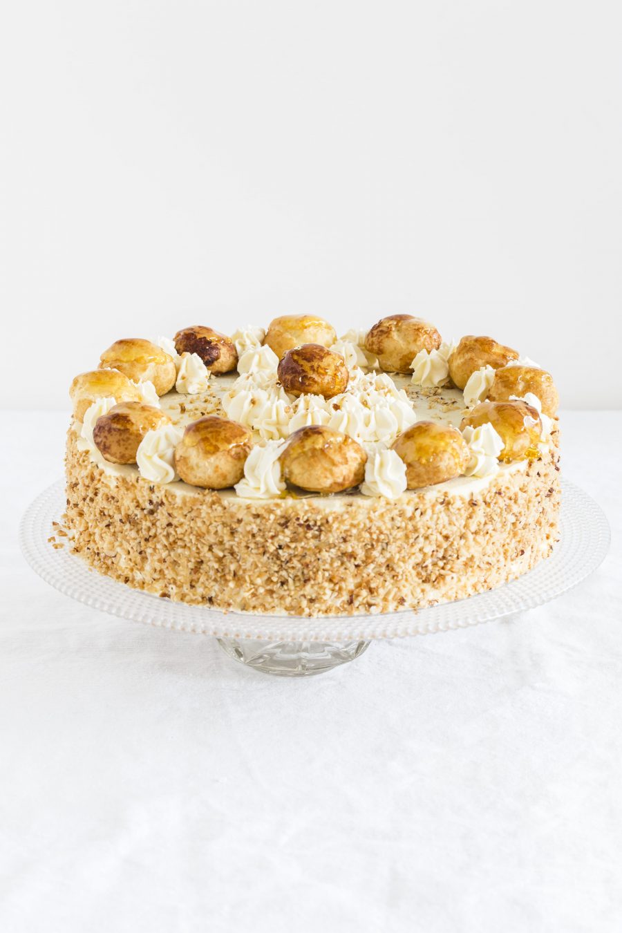 Saint Honorè cake on a cake stand. Choc Crème Patisserie and Vanilla Crème Patisserie, with genoese sponge, puff pastry, surrounded by whipped cream, toasted hazelnuts, and topped with croquembouche choux. Made by Biscotti di Debora, Italian bakers in Cambridge.