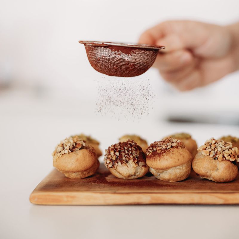Person dusting powdered sugar over petit fours pastries on a wooden board